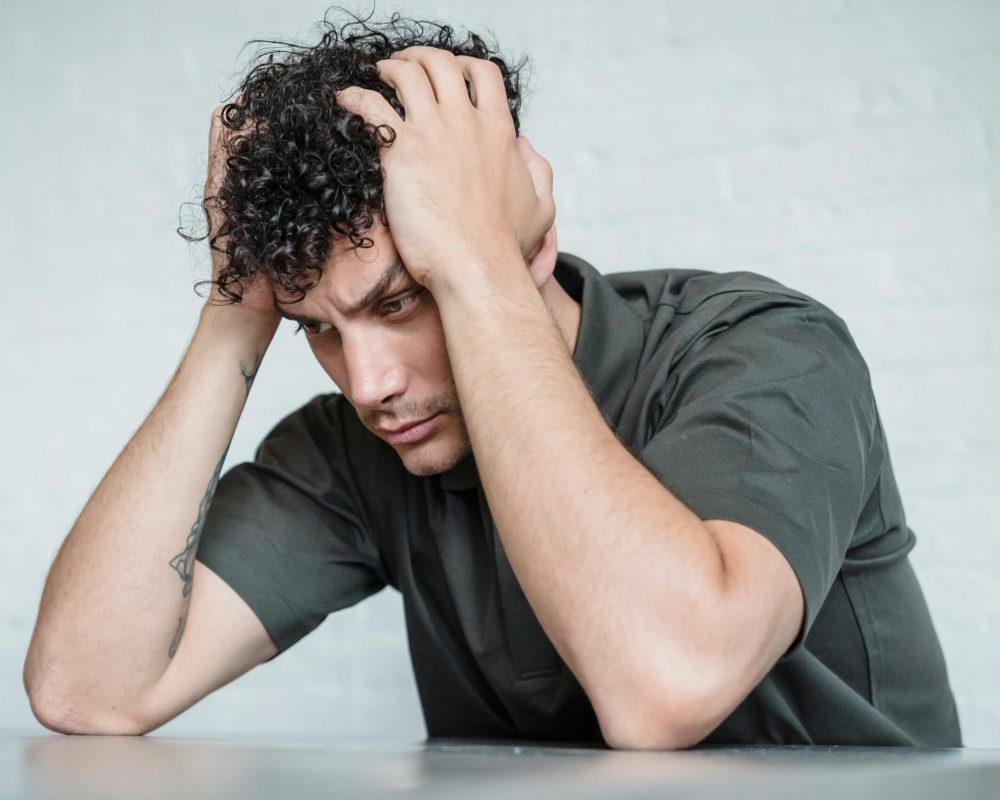 A young man with curly hair looks stressed as he sits with his hands on his head in contemplation.